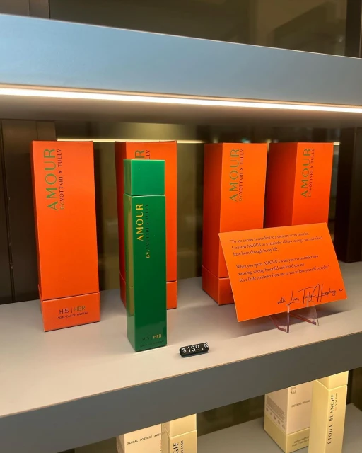 Shelf with green Amour perfume bottle and orange product boxes. Perfume bottle displayed in front of packaging.
