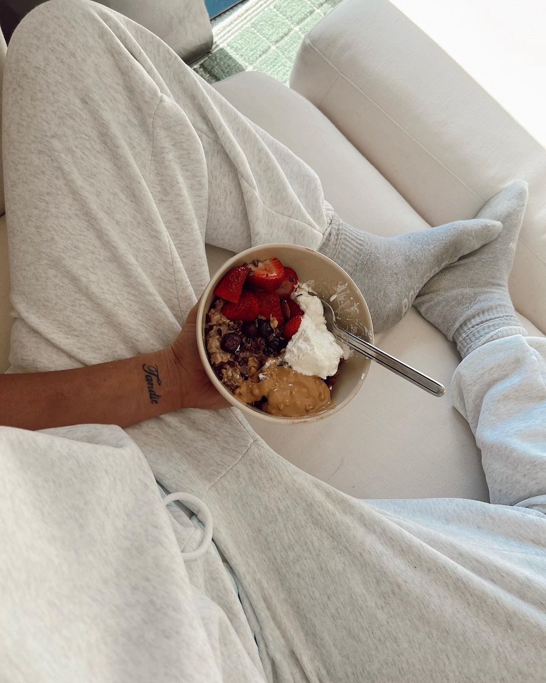 Person in grey sweatpants holding a bowl of food with strawberries, peanut butter, whipped cream and oatmeal while sitting on a couch.