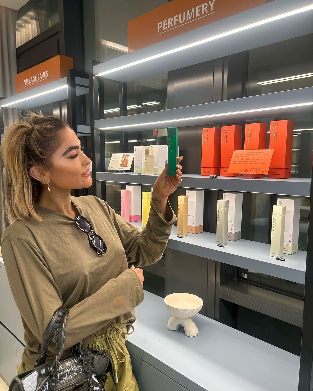 Woman in a perfumery shop holds a green fragrance bottle, surrounded by colorful perfume boxes on illuminated shelves.