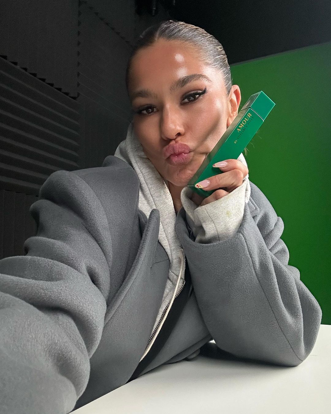 Adult female holds a green perfume bottle indoors, gray hoodie and jacket. Bright green backdrop and diffused lighting.