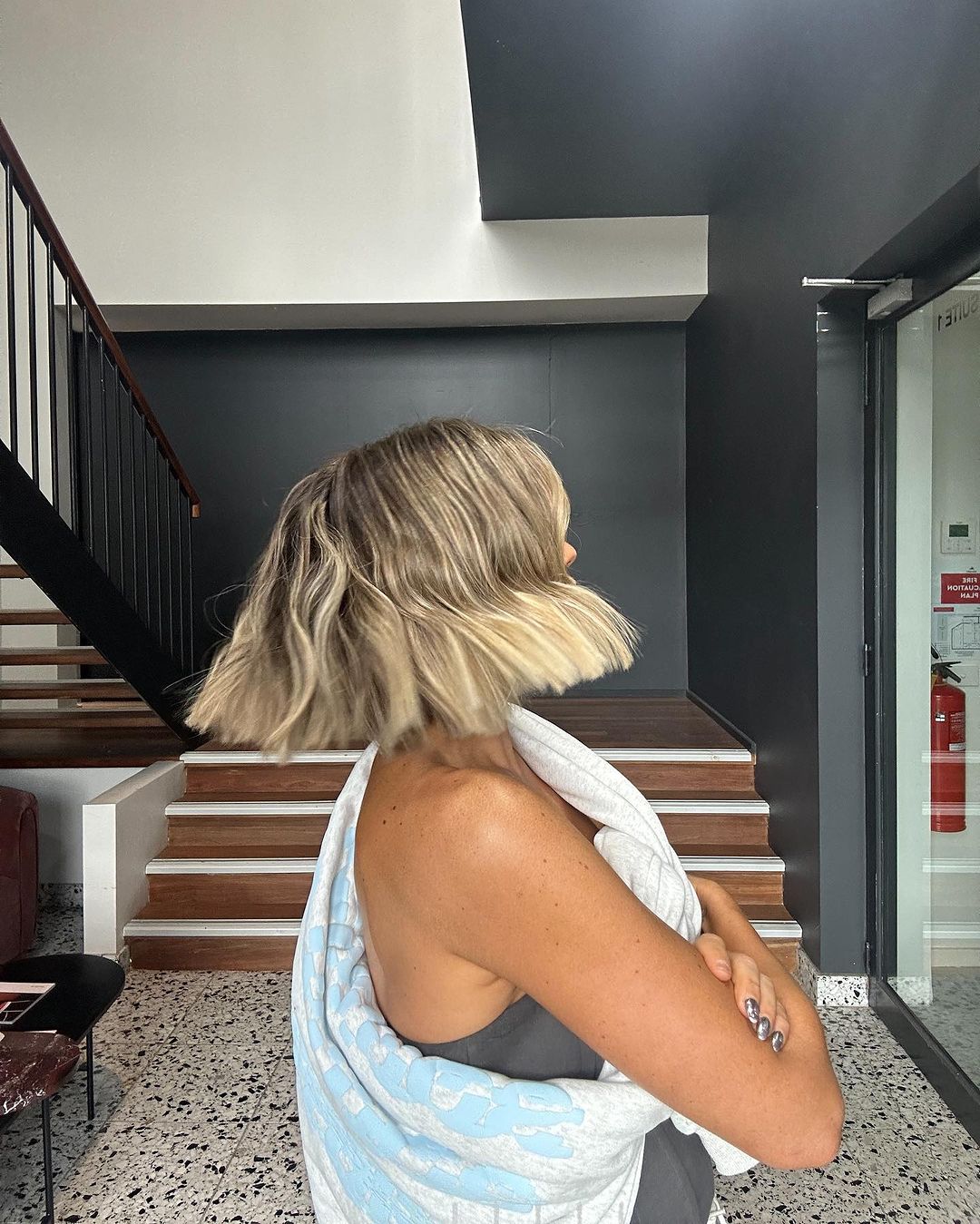 Adult female with blonde bob hairstyle posing indoors near staircase, wearing a grey tank top and a white and blue patterned towel.