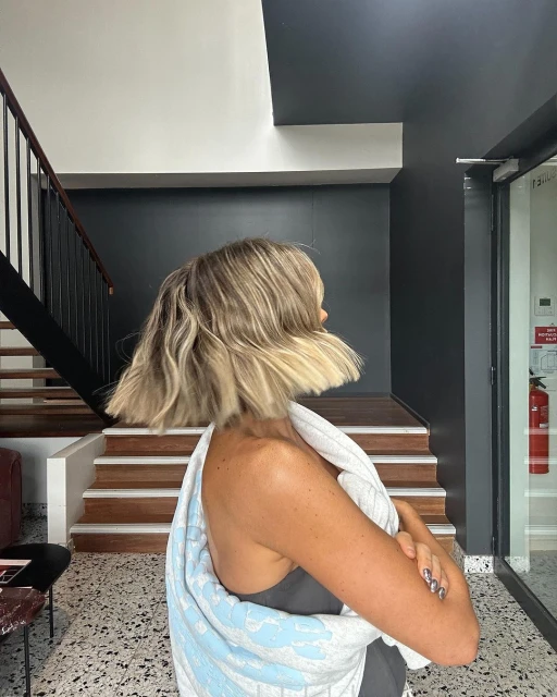 Adult female with blonde bob hairstyle posing indoors near staircase, wearing a grey tank top and a white and blue patterned towel.