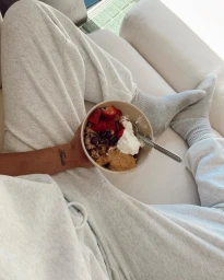 Person wearing grey sweatpants and socks holding a breakfast bowl with oatmeal, fruit, and yogurt