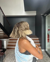 Woman with blonde bob haircut wearing a towel draped over her shoulders in a modern building interior with stairs.