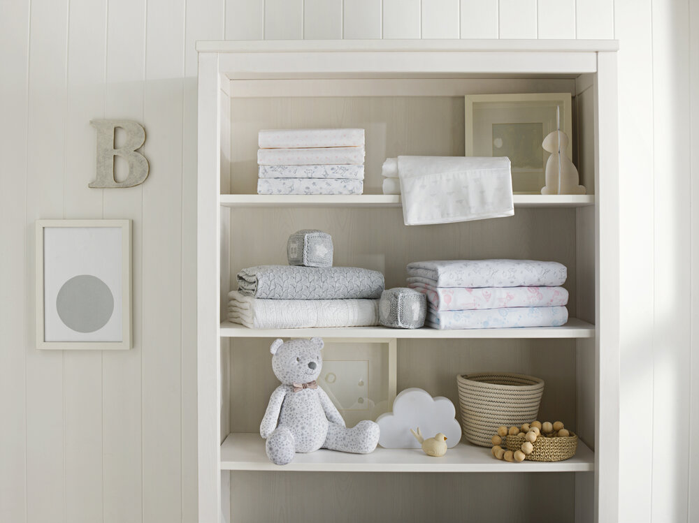 Nursery interior with soft white bookshelf displaying neatly folded linens, a teddy bear, and decorative children's items.