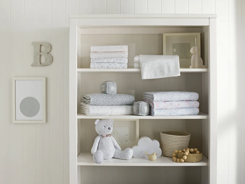 Nursery interior with soft white bookshelf displaying neatly folded linens, a teddy bear, and decorative children's items.