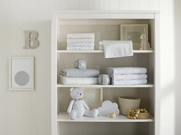 Nursery interior with soft white bookshelf displaying neatly folded linens, a teddy bear, and decorative children's items.