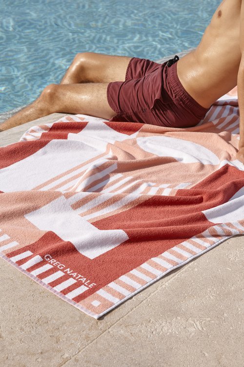 Man in swim trunks rests poolside on an orange, peach, and white geometric beach towel by Greg Natale in bright sunlight.