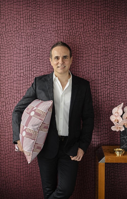 Adult male in a black blazer and dark pants, holding a patterned pillow, stands indoors against a textured wallpapered wall.