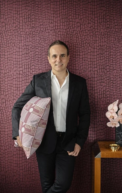 Adult male in a black blazer and dark pants, holding a patterned pillow, stands indoors against a textured wallpapered wall.