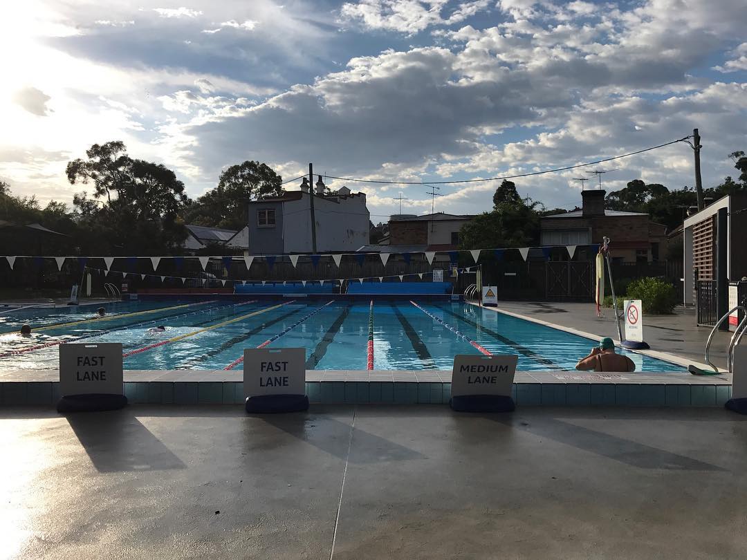 Outdoor swimming pool with lane markers and buoys on a partly cloudy day, featuring 'Fast Lane' and 'Medium Lane' signage.