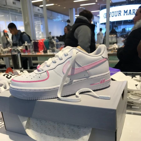 White leather Nike Air Force 1 sneaker with pink details sitting on a gray box in a brightly lit indoor location with people.
