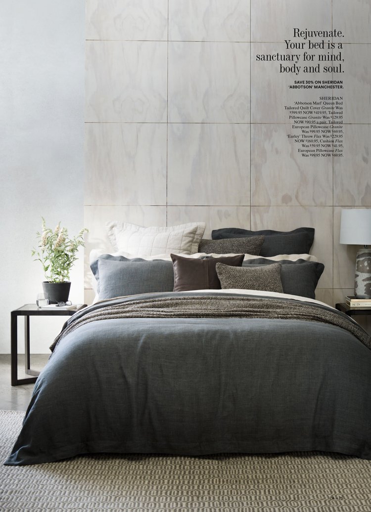 Bedroom interior featuring a grey duvet cover on a bed with multiple pillows, a side table with a lamp, and a potted plant.