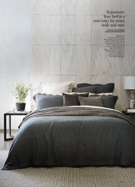 Bedroom interior featuring a grey duvet cover on a bed with multiple pillows, a side table with a lamp, and a potted plant.