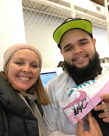 Two people pose indoors with a customized Nike Air Force 1 shoe featuring pink and black accents, held by the person on the right.