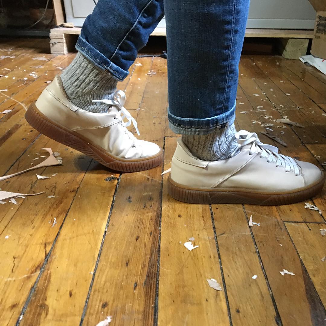 Person stands on a wooden floor wearing jeans, gray socks, and white sneakers with brown soles, with scraps of paper scattered nearby.