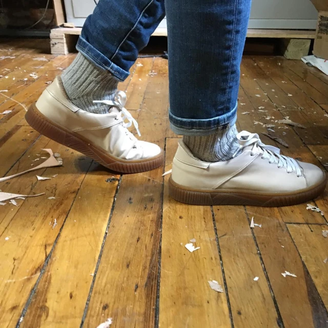 Person stands on a wooden floor wearing jeans, gray socks, and white sneakers with brown soles, with scraps of paper scattered nearby.