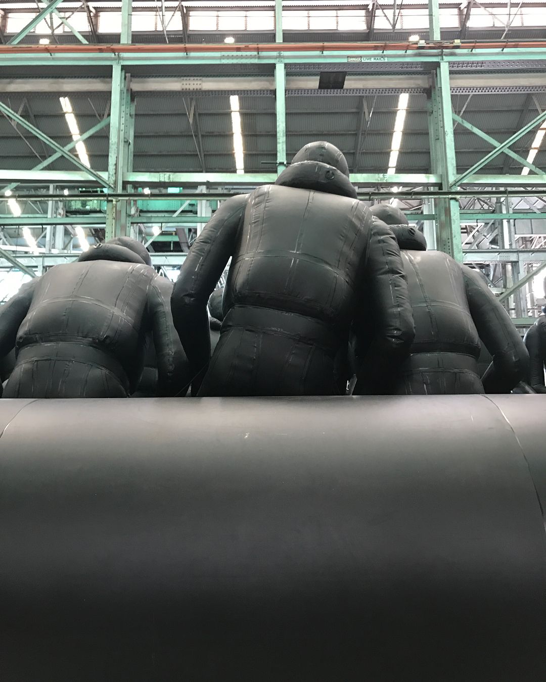 A group of figures in puffy black suits sit in an industrial space under metal girders and bright lights from an eye-level angle.