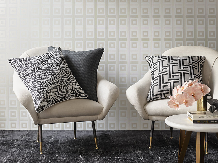 Interior design featuring two cream-colored chairs with black patterned pillows, a marble side table with books, and a gold vase with flowers.