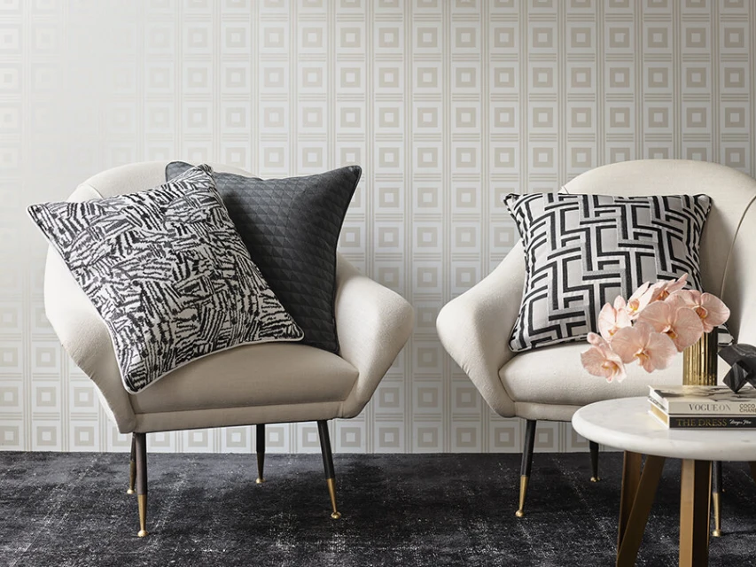 Interior design featuring two cream-colored chairs with black patterned pillows, a marble side table with books, and a gold vase with flowers.
