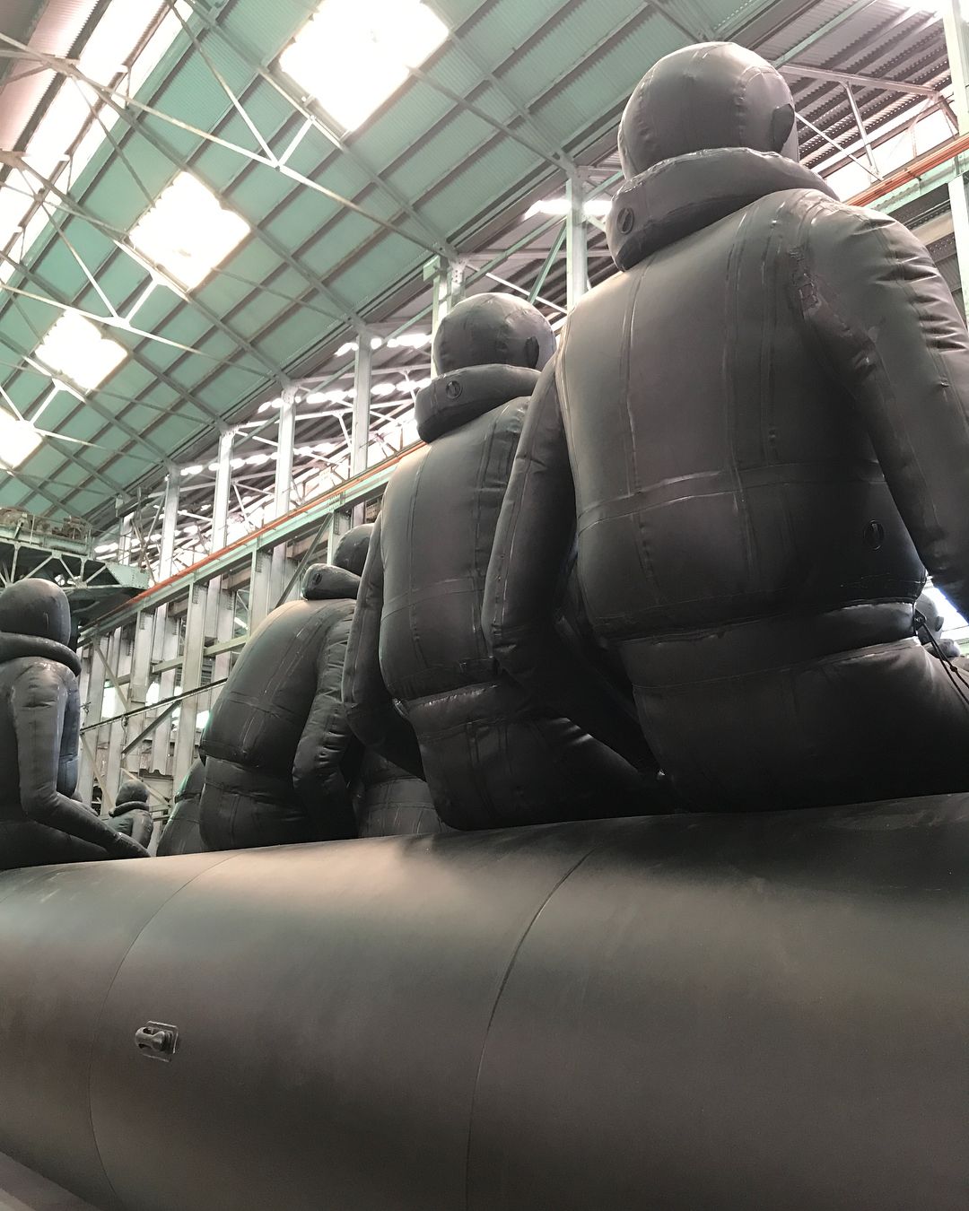 Row of inflated, human-shaped black figures sitting atop an inflated black raft or tube in an industrial building with a high ceiling.