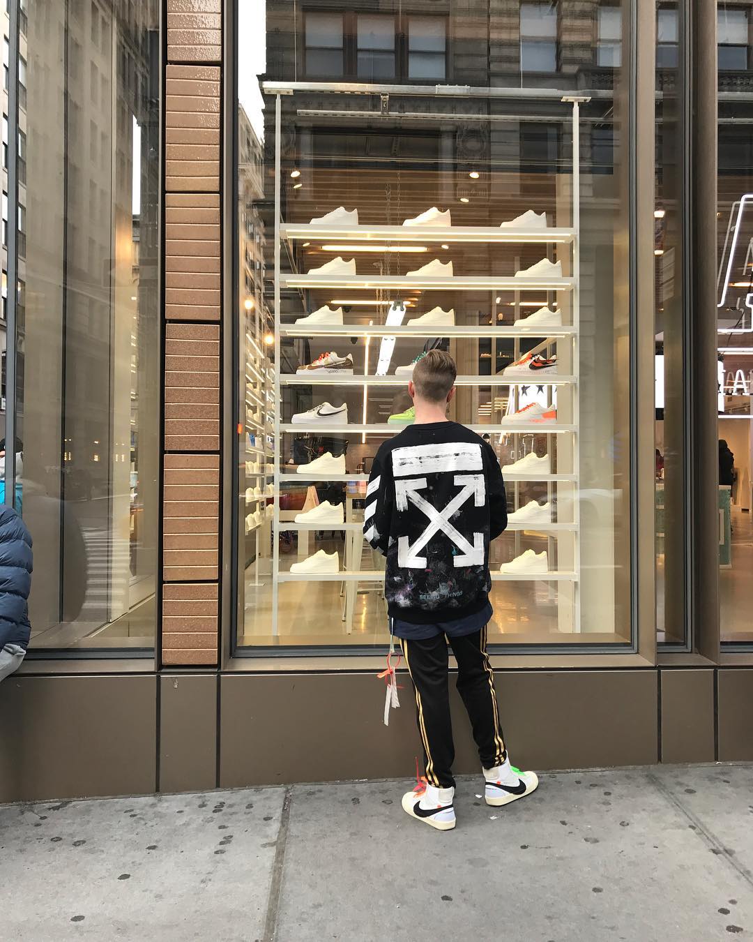 Man in Off-White jacket looks at athletic shoes in a retail storefront display window in a city environment on a cloudy day.