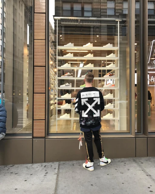 Man in Off-White jacket looks at athletic shoes in a retail storefront display window in a city environment on a cloudy day.
