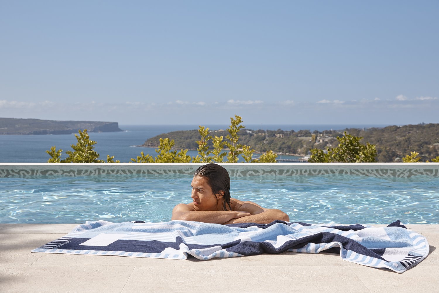 A man in a pool with a patterned beach towel, looking out at a coastal view under a blue sky on a bright, sunny day.
