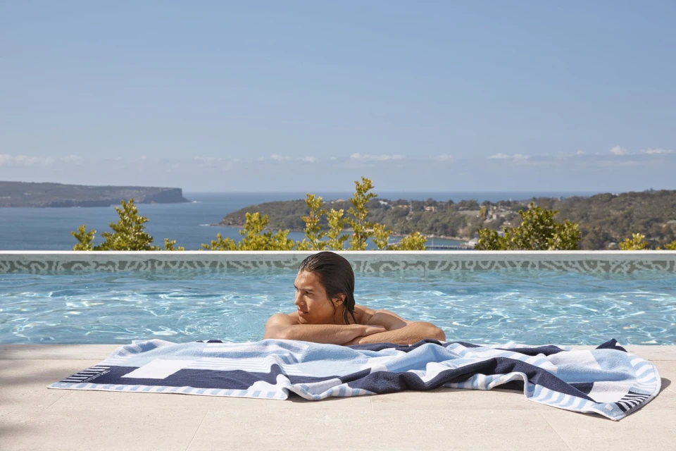 A man in a pool with a patterned beach towel, looking out at a coastal view under a blue sky on a bright, sunny day.