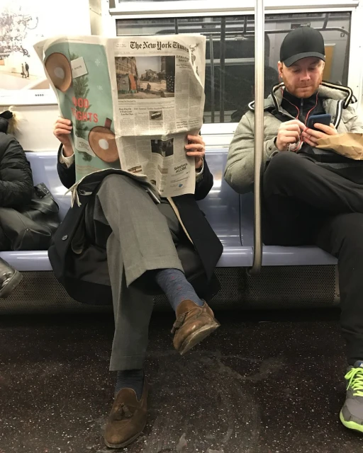 Man reading The New York Times newspaper on the subway while another man uses his smartphone nearby in an indoor setting.