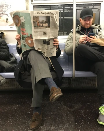 Man reading The New York Times newspaper on the subway while another man uses his smartphone nearby in an indoor setting.