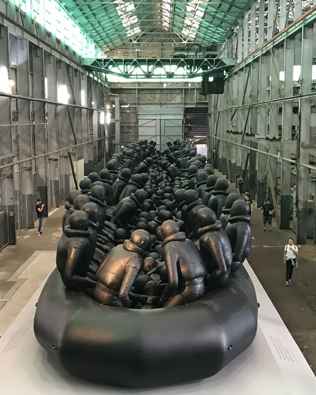 Indoor view of a large art installation with a black inflatable boat crowded with seated humanoid figures in a warehouse space.