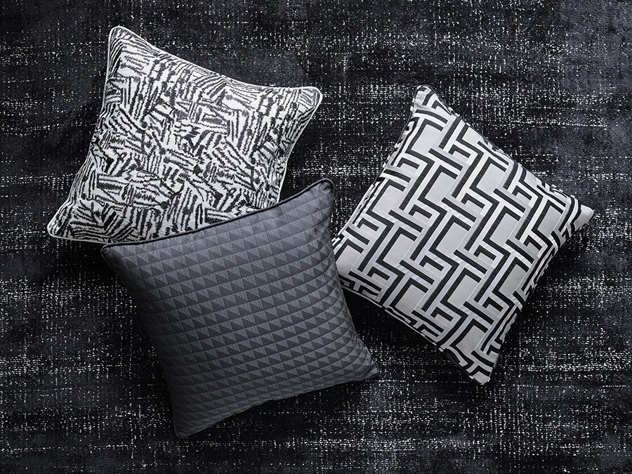 Three decorative pillows with black and white geometric patterns are arranged on a textured black rug in an indoor setting.