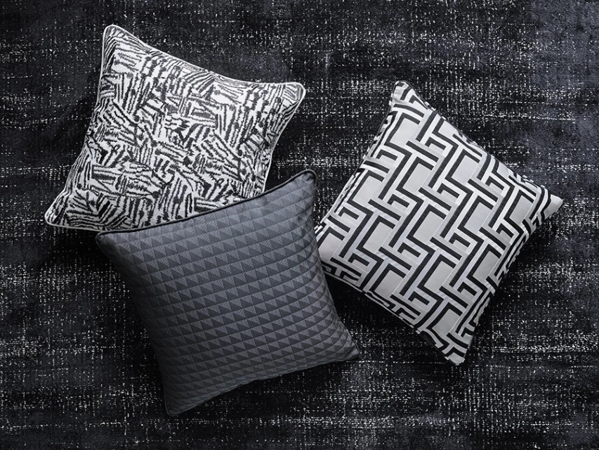 Three decorative pillows with black and white geometric patterns are arranged on a textured black rug in an indoor setting.