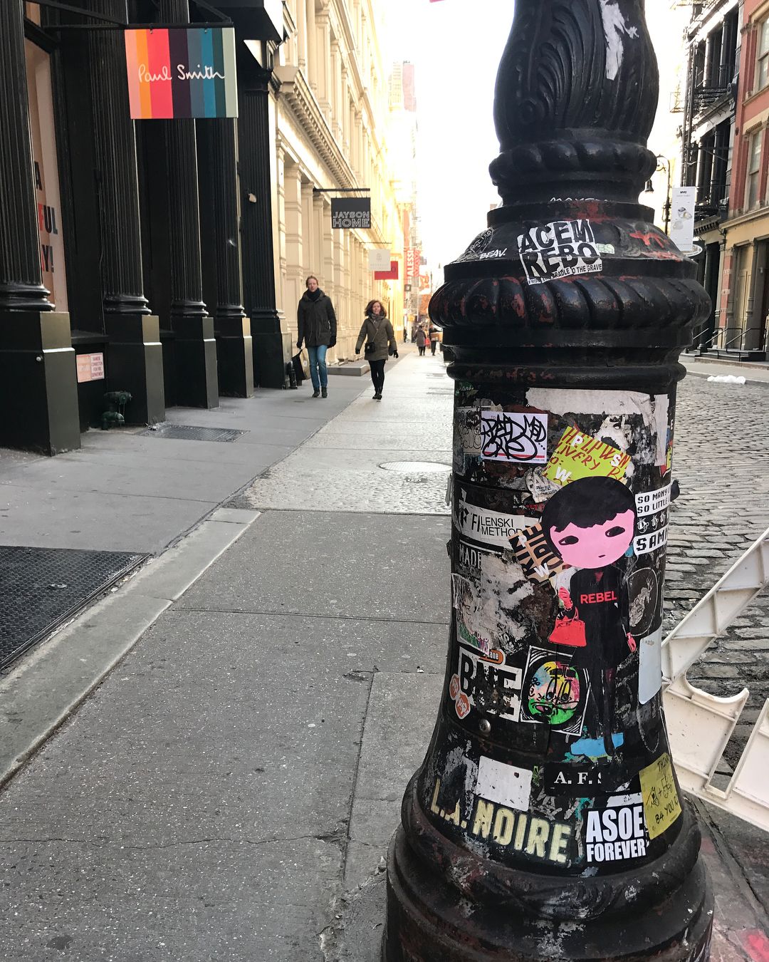Street scene with two pedestrians walking past stores and a sticker-covered pole with posters, offering an urban, artistic vibe.