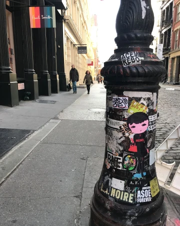 Street scene with two pedestrians walking past stores and a sticker-covered pole with posters, offering an urban, artistic vibe.
