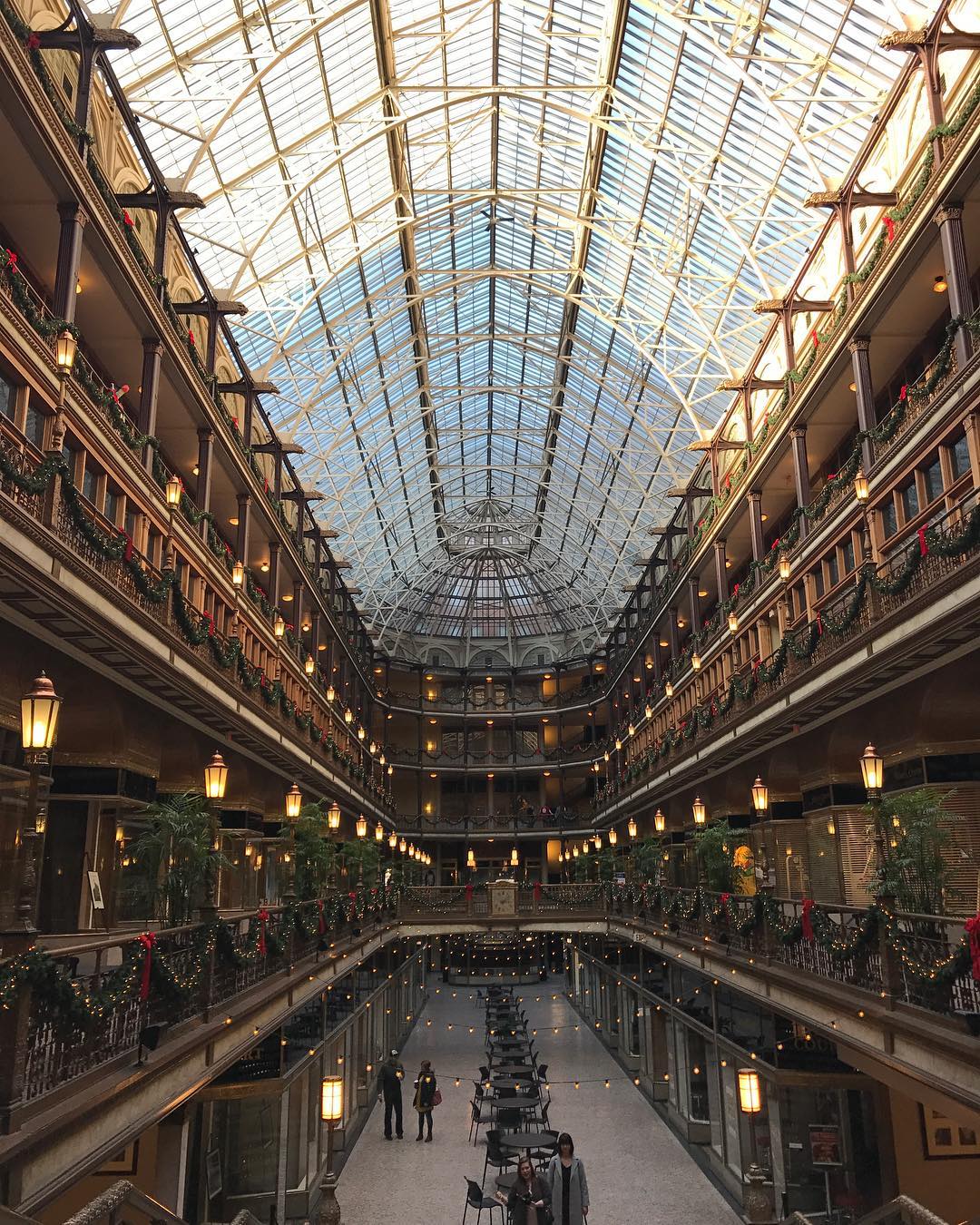 Interior view of a grand atrium with a glass ceiling, multiple levels with ornate railings, string lights, and people walking.