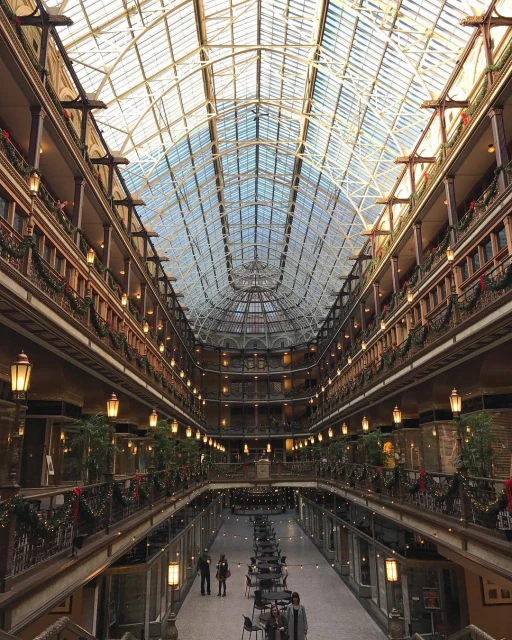 Interior view of a grand atrium with a glass ceiling, multiple levels with ornate railings, string lights, and people walking.