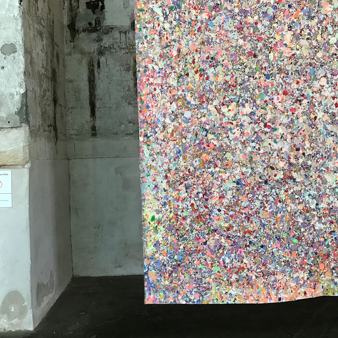 Abstract art installation featuring colorful speckled pattern in shades of pink, purple, and white alongside weathered concrete wall.