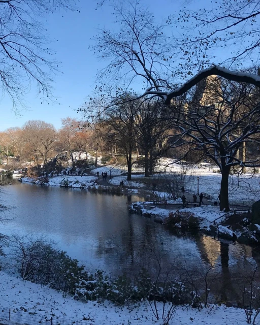 Snowy winter landscape featuring a serene lake, bare trees, and a clear blue sky. People walk along a path in the distance.