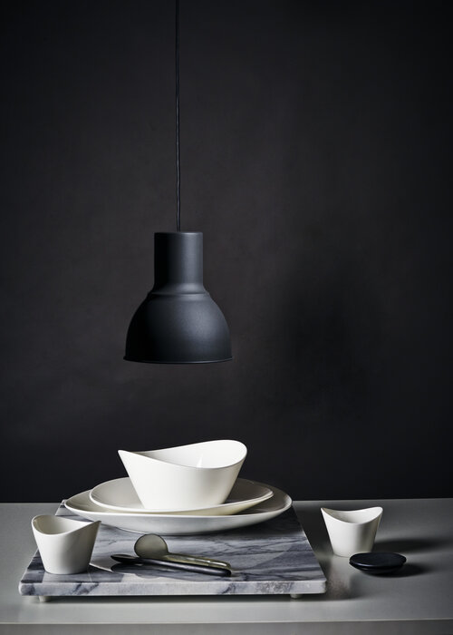 Still life photograph of white ceramic dishes and a gray marble cutting board under a modern black pendant lamp against a dark backdrop.