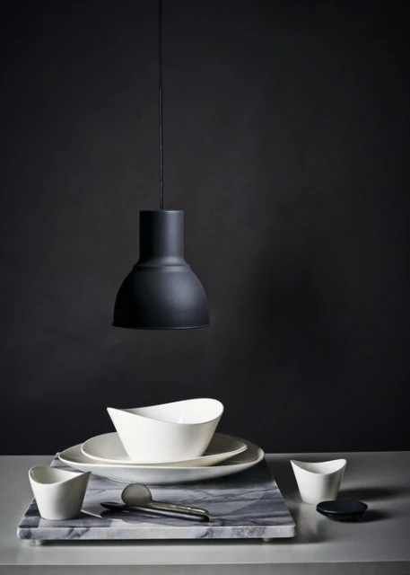 Still life photograph of white ceramic dishes and a gray marble cutting board under a modern black pendant lamp against a dark backdrop.