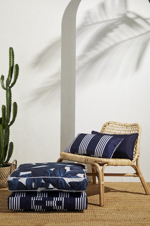 Stack of cushions, a woven chair with pillows, a cactus in a basket, with a palm leaf shadow on a white wall.
