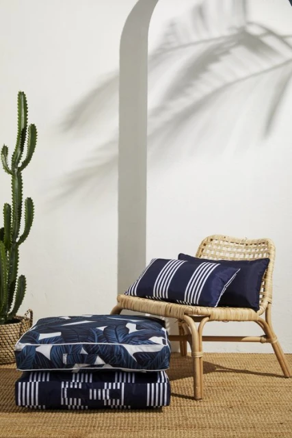 Stack of cushions, a woven chair with pillows, a cactus in a basket, with a palm leaf shadow on a white wall.