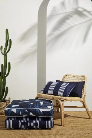 Stack of cushions, a woven chair with pillows, a cactus in a basket, with a palm leaf shadow on a white wall.