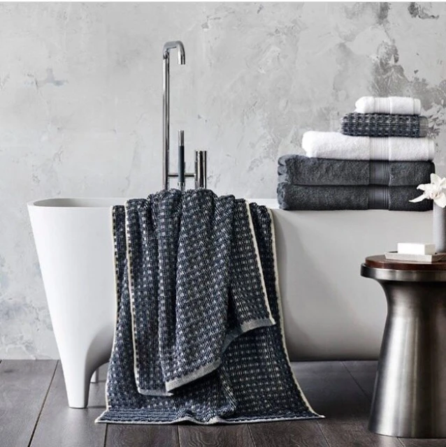 Elegant bathroom interior featuring a freestanding white bathtub, chrome faucet, grey patterned towel, stacked towels, and a modern side table.