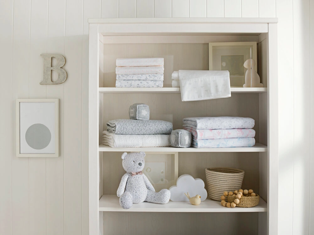 White wooden bookshelf styled with folded baby bedding, a grey teddy bear, knitted cubes, a basket, and framed artwork