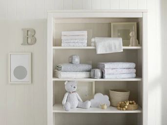 White wooden bookshelf styled with folded baby bedding, a grey teddy bear, knitted cubes, a basket, and framed artwork