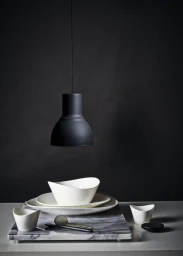 Minimalist tabletop still life featuring white ceramic bowls, plates, dark spoons, and a black pendant lamp against a dark textured background.