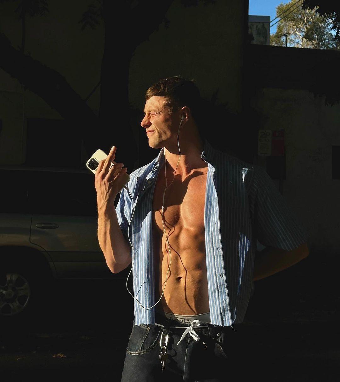 Attractive man with muscular build wearing blue shirt and jeans, holding phone in sunlight in urban environment with a car in background.
